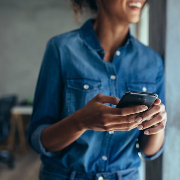 Woman in casuals standing in office by a window with phone in hand.