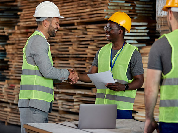 Smiling business partners in safety gear shaking hands at a lumber facility